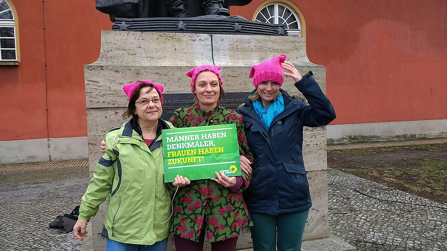 drei Frauen mit pinken Mützen, die die Katzenohren der "Pussycats" tragen, posieren vor dem Sockel eines Denkmals. Das sieht man nicht, aber es ist General Friedrich Wilhelm von Steuben, den die Grünen ausgewählt haben. Es ist kalt und windig an diesem Märztag, dem Weltfrauentag.