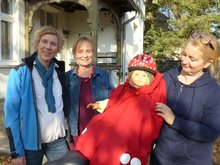 Dilassi-Chefin Anne Laßhofer (links), die Brandenburger Landesvorsitzende Petra Budke (mittig), Sprecherin des Ortsverbandes Falkensse Yvonne Scherzer (rechts) mit dem WichtelWarm an Demo-Puppe Emma-Susi. Die drei Frauen stehen bei schönem Herbstwetter draußen um das Fahrrad mit der lebensgroßen Puppe auf dem Rücksitz herum, der WichtelWarm leuchtet rot in der Herbstsonne. Die Gründerin trägt Outdoor-Kleidung, praktisch angezogen, und wirkt unter dem struwweligen kurzen Blondschopf sehr fröhlich.