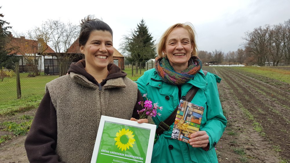 Grüne Gründerin Nina Keller auf ihrem Feld mit der Landesvorsitzenden Petra Budke Man sieht zwei lachende Frauen im Herbst, der Hintergrund ist kahl, das Feld dahinter abgeerntet. Nur vereinzelt blühen noch lila die Kartäusernelken. Links mit dunklen Haaren ist Nina Keller, die Botanikerin und Gründerin von "Die Wildblume". Rechts mit der Urkunde in der Hand ist die Landesvorsitzende von Bündnis 90/Die Grünen in Brandenburg, Petra Budke. Um sie herum sind viele Journalisten, die man aber auf dem Bild nicht sehen kann.