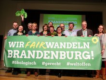 Auf einer Bühne stehen viele lachende Menschen: es sind die frisch gewählten Kandidatinnen und Kandidaten auf dem Landesparteitag in Fürstenwalde. Sie stehen auf der Bühne im Saal des Fürstenwalder Hofes und halten ein Banner in der Hand mit der Aufschrift: Wir fairwandeln Brandenburg - ökologisch, gerecht, weltoffen. Das ist das Motto des Parteitags und des anstehenden Wahlkampfes im Superwahljahr 2019. Das "Fairwandeln" ist natürlich ein Wortspiel für "fair" und "verwandeln".
