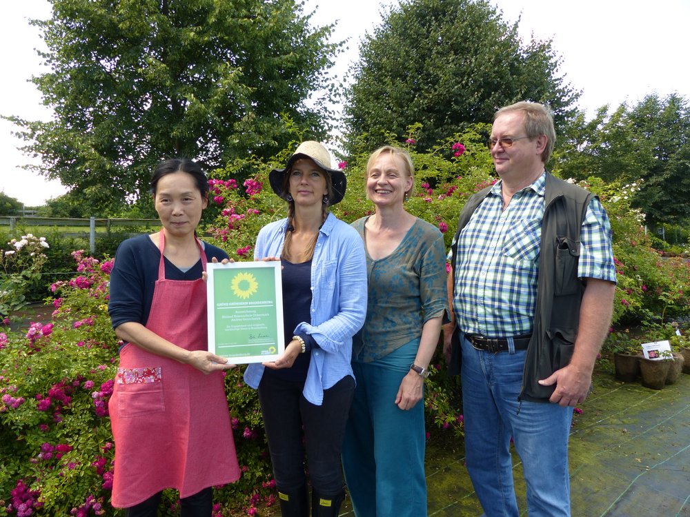 Auf dem Foto vor vielen pinkfarbenen kleinen Rosenblüten posieren vier Menschen: links die japanische Köchin des Rosen Cafés Akiko Hashimoto, daneben die Grüne Gründerin Andrea Genschorek, daneben die Landesvorsitzende der Brandenburger Bündnisgrünen, Petra Budke, und ganz rechts Thomas Dyhr, der als Direktkandidat für die Bundestagswahl 2017 für die Uckermark antritt. Frau Genschorek hält die Urkunde in der Hand, sie trägt einen Sonnenhut aus Stroh. Frau Hashimoto hat eine pinkfarbene Küchenschürze um. Es wirkt harmonisch an diesem sonnigen Tag im Juli.