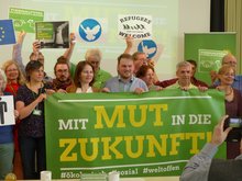 Viele Menschen aus dem Landesvorstand und die Direktkandidat*innen halten ein Transparent auf einer Bühne, darauf steht: Mit Mut in die Zukunft - #ökologisch #sozial # weltoffen. Es ist im Rathaus Kleinmachnow am 1. April 2017 beim Landesparteitag aufgenommen. Die Menschen halten Piktogramme hoch, z.B. mit der Friedenstaube oder mit "Refugees welcome".