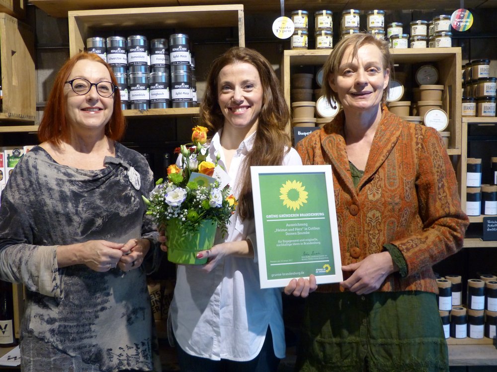 Drei Frauen strahlen in die Kamera und stehen vor einem Regal voller regionaler Lebensmittel. Es sind von BÜNDNIS 90/DIE GRÜNEN die Kreisvorsitzende Cottbus, Petra Weissflog, und die Brandenburger Landesvorsitzende Petra Budke. Sie halten für die stolze Inhaberin des Cafés "Heimat und Herz ein Blumengesteck und eine Urkunde in der Hand.