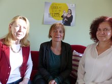 von links nach rechts sitzen auf einem roten Sofa im Geburtshaus Am Neuen Garten: Martina Schulze, die 1. Vorsitzende des Hebammenverbands Brandenburg, die Landesvorsitzende Petra Budke in der Mitte, rechts die Hebamme Peggy Jahnel. Hinter ihnen an der Wand hängt ein Poster mit einer Mutter und ihrem Baby auf dem Arm, dazu die Aufschrift: Geboren mit der Hilfe ihrer Hebamme.
