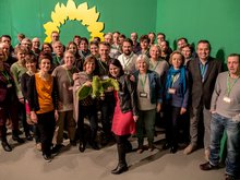 Ein Gruppenbild vor dem grünen Hintergrund mit Sonnenblume. Mindestens 40,50 Menschen, es sind die Delegierten und Fans aus Brandenburg auf dem Bundesparteitag in Hannover. Davor eine sichtlich jubelnde, bewegte Annalena Baerbock mit dem Maskottchen "Gudrun Plüschadler". Auch Robert Habeck ist auf dem Bild zu sehen, der sich die Doppelspitze mit ihr teilen wird.