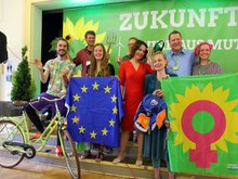 Vor dem Banner "Zukunft wird aus Mut gemacht" posieren Annalena Baerbock, Petra Budke, Clemens Rostock, Direktkandidaten und ein neues Mitglied mit symbolischen Gegenständen. Z.B. einem Schulranzen für Bildung, einem Windrad für Erneuerbare, Energien, eine Milchflasche mit verdrecktem original aus der Spree genommenem Wasser für eine saubere Spree, eine Forke für gesundes Essen und gute Landwirtschaft. Lachende Gesichter im vollen Saal.