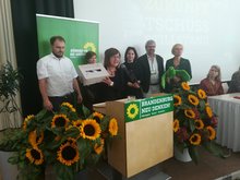 Auf der Bühne: Ursula Nonnemacher hält eine Box hoch, darin konnten die Mitglieder Anregungen und Vorschläge für die zukünftigen Verhandlungen für eine Koalition einwerfen. Die Mitglieder des Landesparteirats bedanken sich bei den Delegierten auf dem Kleinen Parteitag in Kleinmachnow. Um Frau Nonnemacher herum stehen - sichtlich erschöpft - Clemens Rostock, Annalena Baerbock, Petra Budke, Julia Schmidt, Axel Vogel.